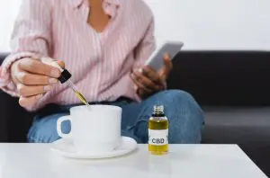 femme afro-américaine tenant un smartphone et une pipette avec du liquide CBD près d'une tasse